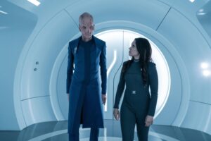 A tall, thin alien and a human woman walk through the tunnel of a spaceship.